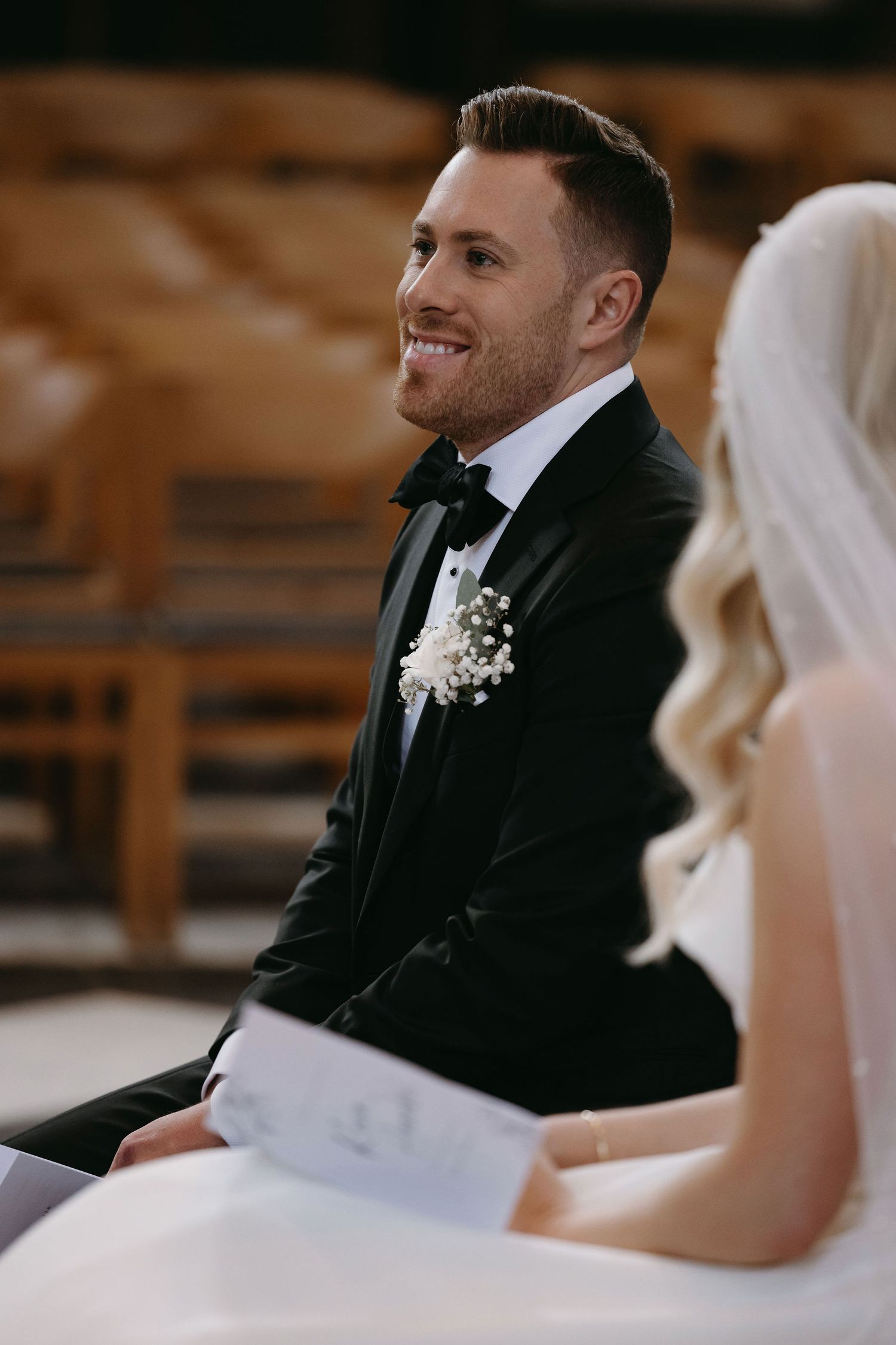 A groom in classic black tuxedo smiles during wedding ceremony.