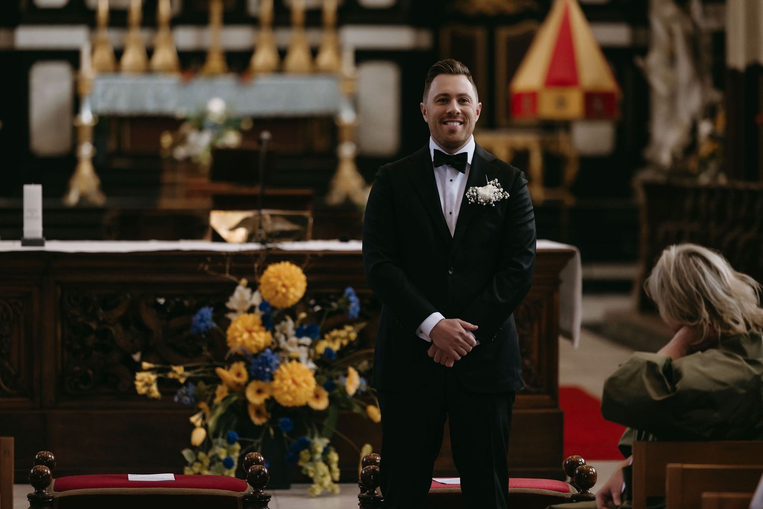 Groom in classic black tuxedo waiting at altar with yellow floral arrangements in church setting.