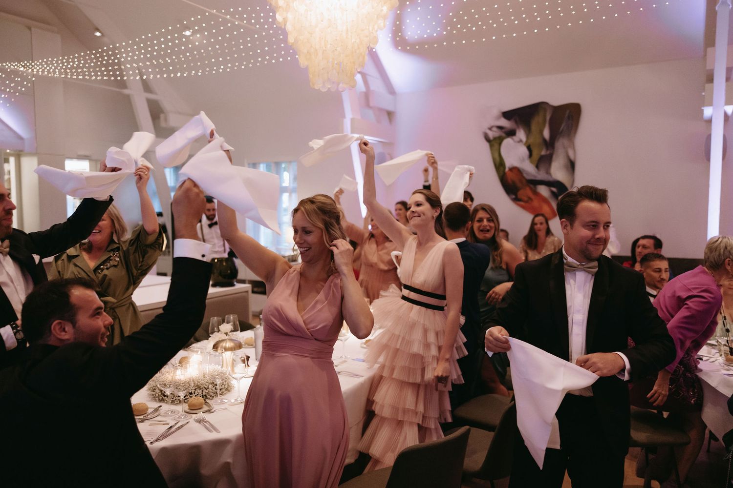 Wedding reception guests celebrate with napkins in a festive ballroom under elegant chandeliers.