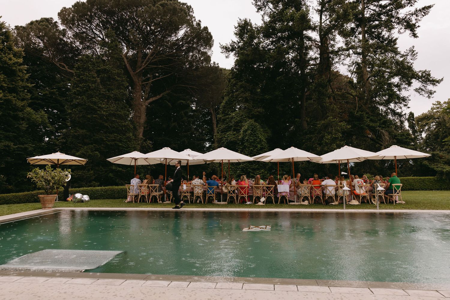 Panoramic view of outdoor pool area with umbrellas and wedding guests gathered nearby.