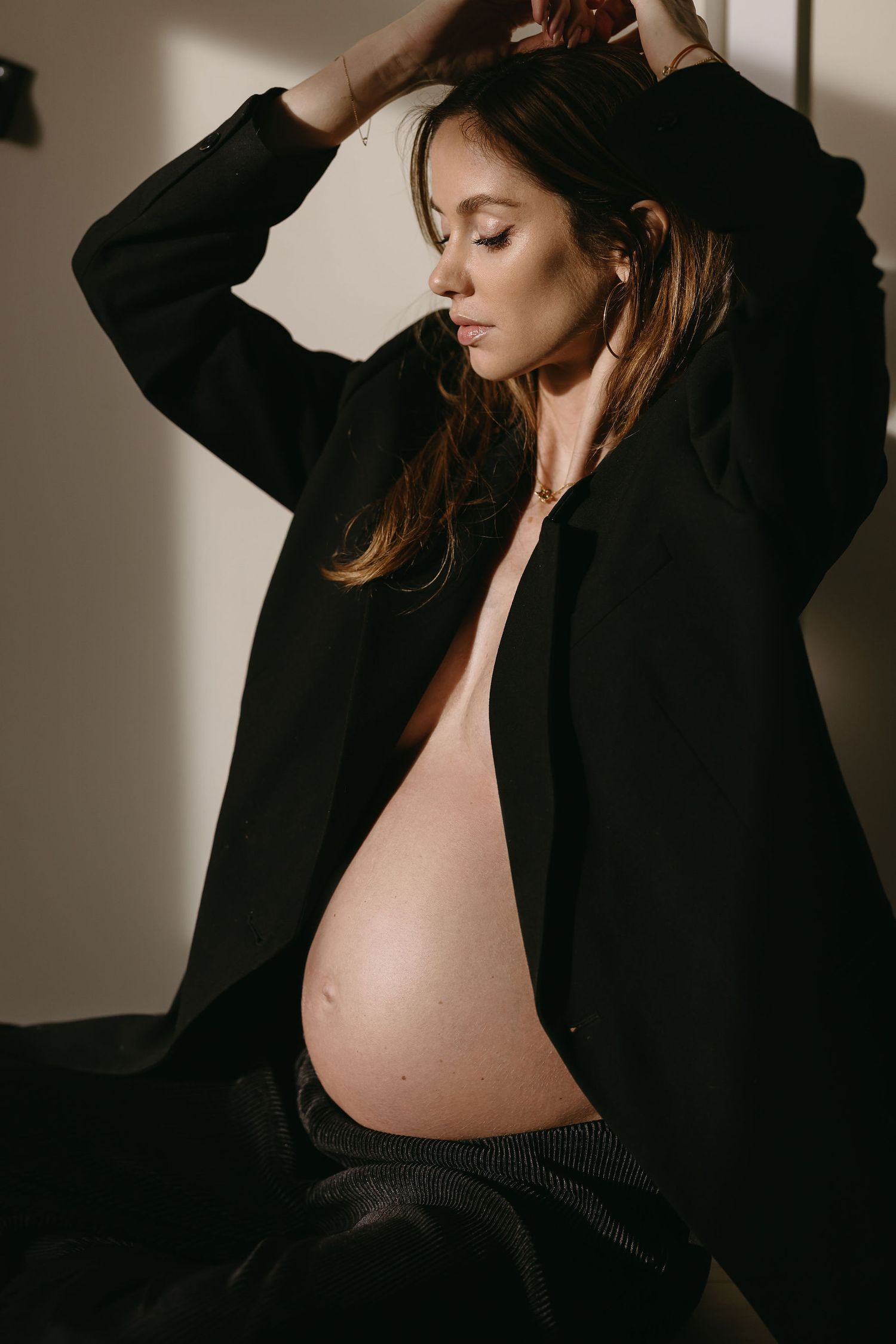 Artistic maternity portrait in black coat showing pregnant belly against moody lighting.