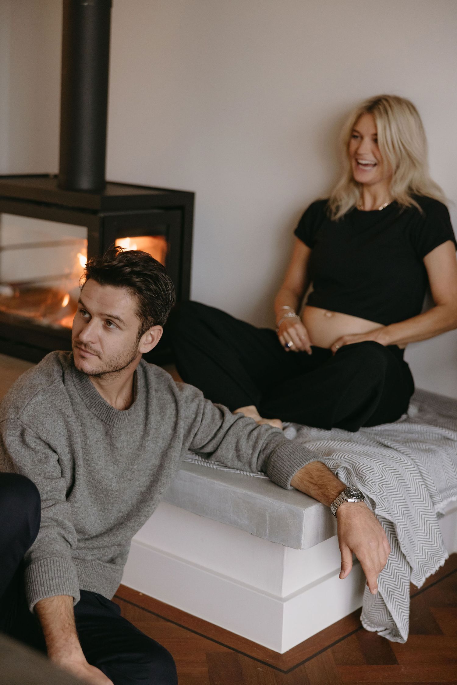 Casual maternity photo of couple lounging by fireplace in cozy home setting.
