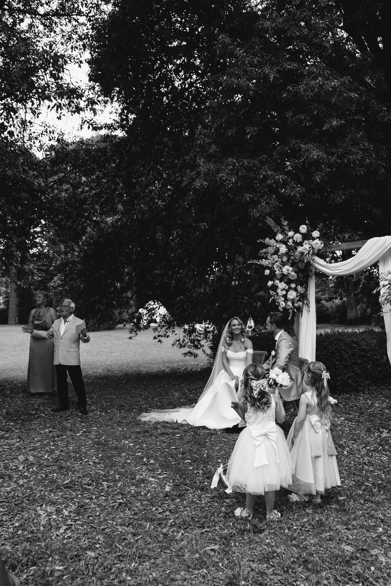Outdoor garden wedding ceremony with white chairs and floral arrangements under trees.