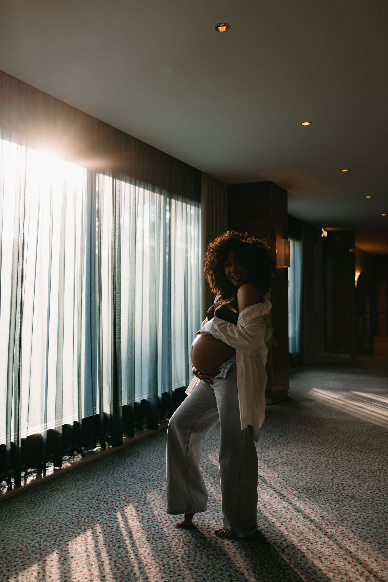Backlit maternity silhouette standing by floor-to-ceiling windows.
