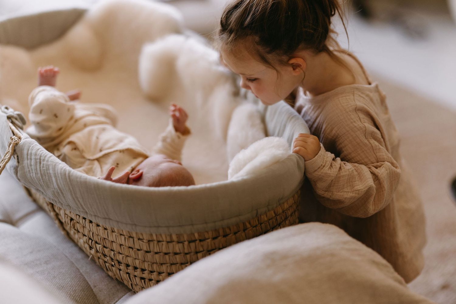 A child peering into a woven basket containing a sleeping baby on white bedding.