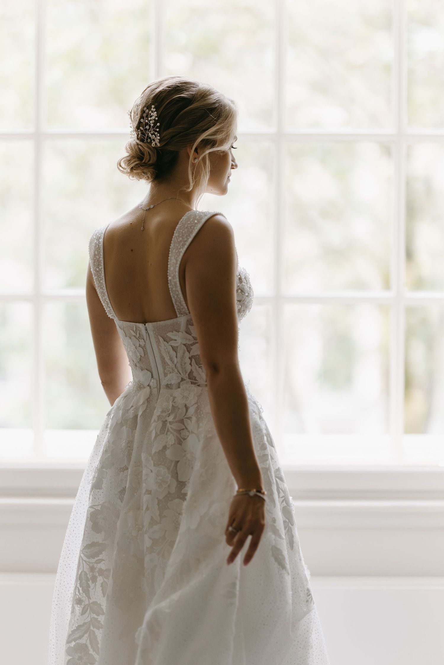 Back view of bride in detailed lace wedding dress standing by window in soft natural light.