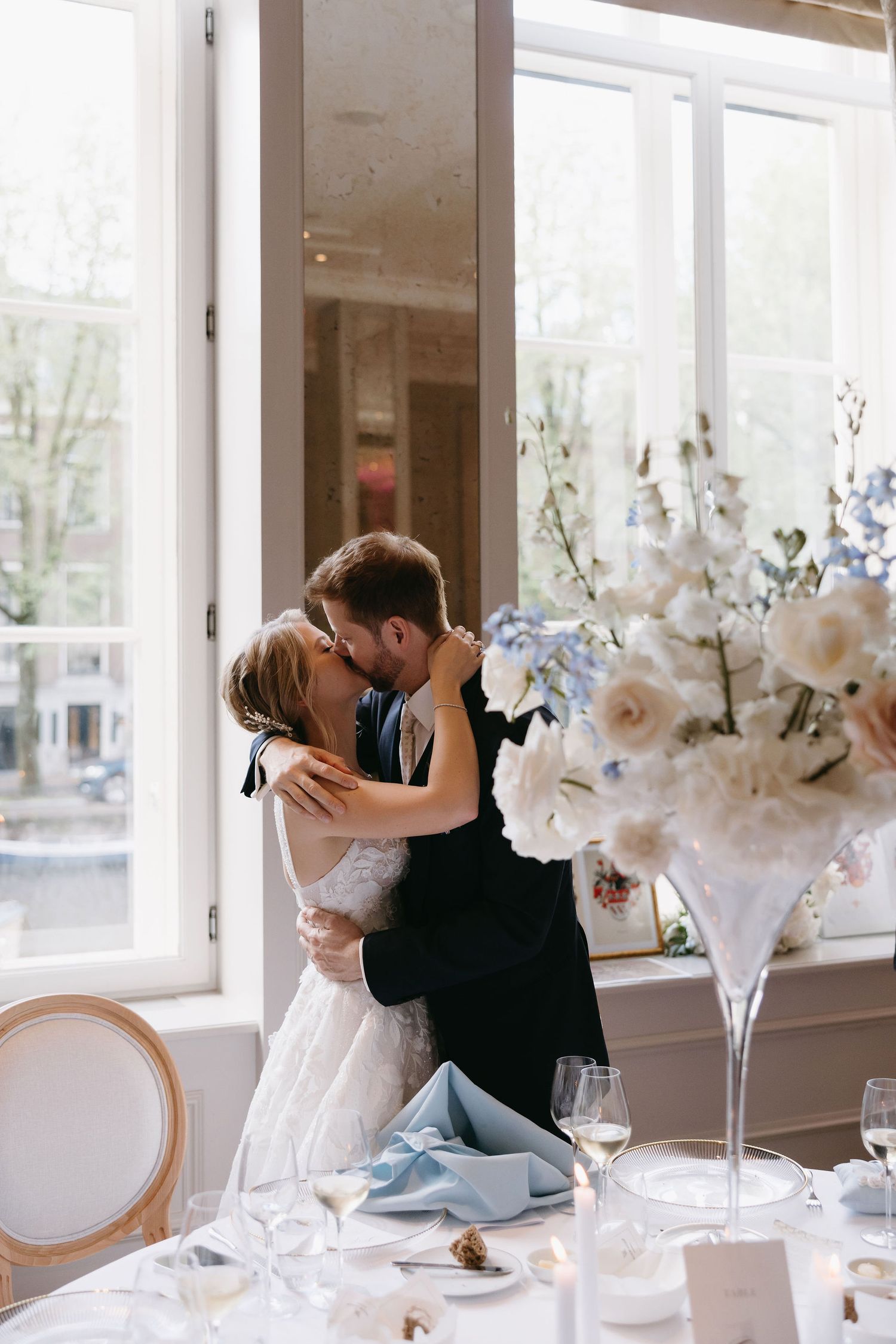 Romantic moment between couple at elegant reception with white floral centerpieces and bright windows.