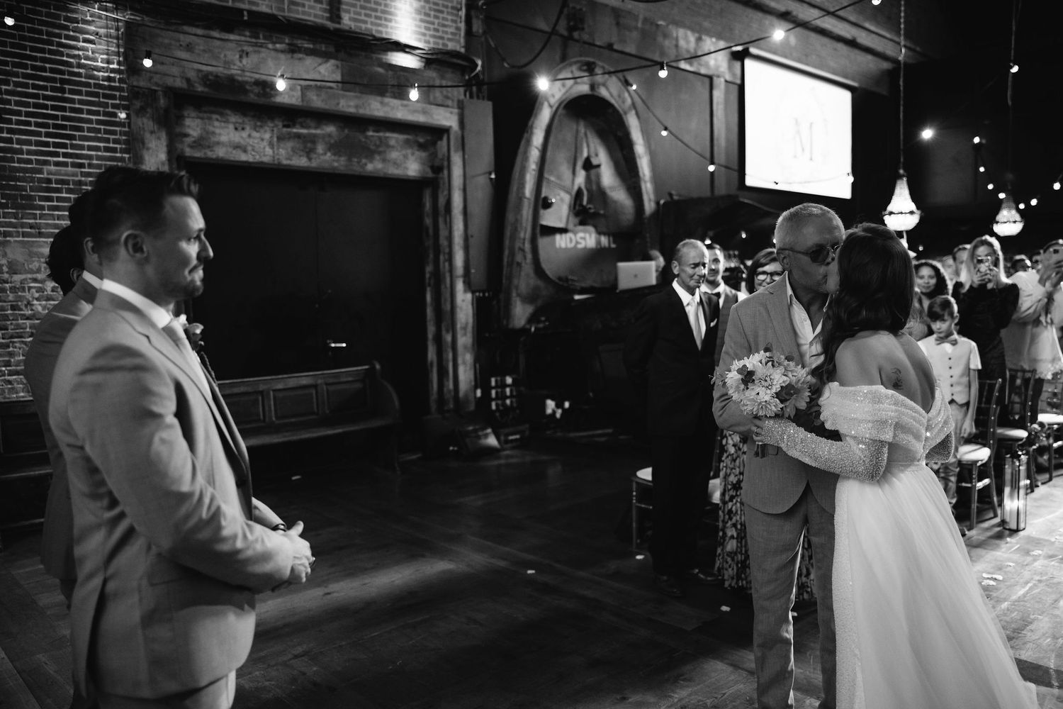 Black and white photo of wedding guests mingling in an industrial-style reception venue.