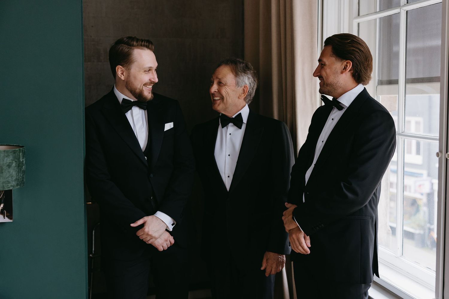 Groomsmen in tuxedos gathered by window for pre-ceremony conversation.