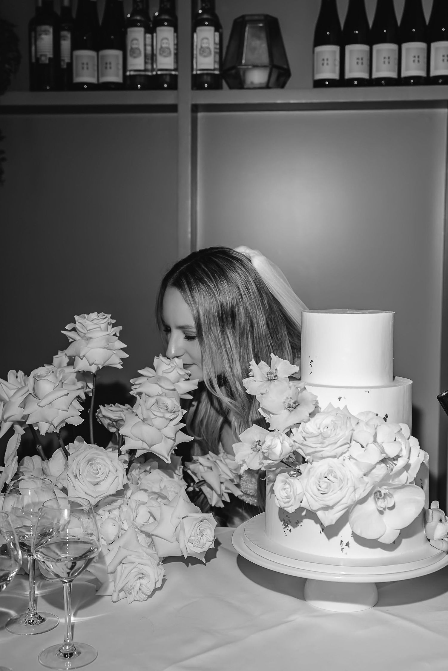 An elegant white wedding cake decorated with roses sits next to floral arrangements.