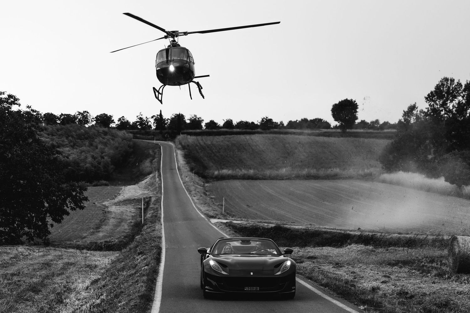 A dramatic black and white photo of a helicopter hovering over a sports car on an empty rural road.