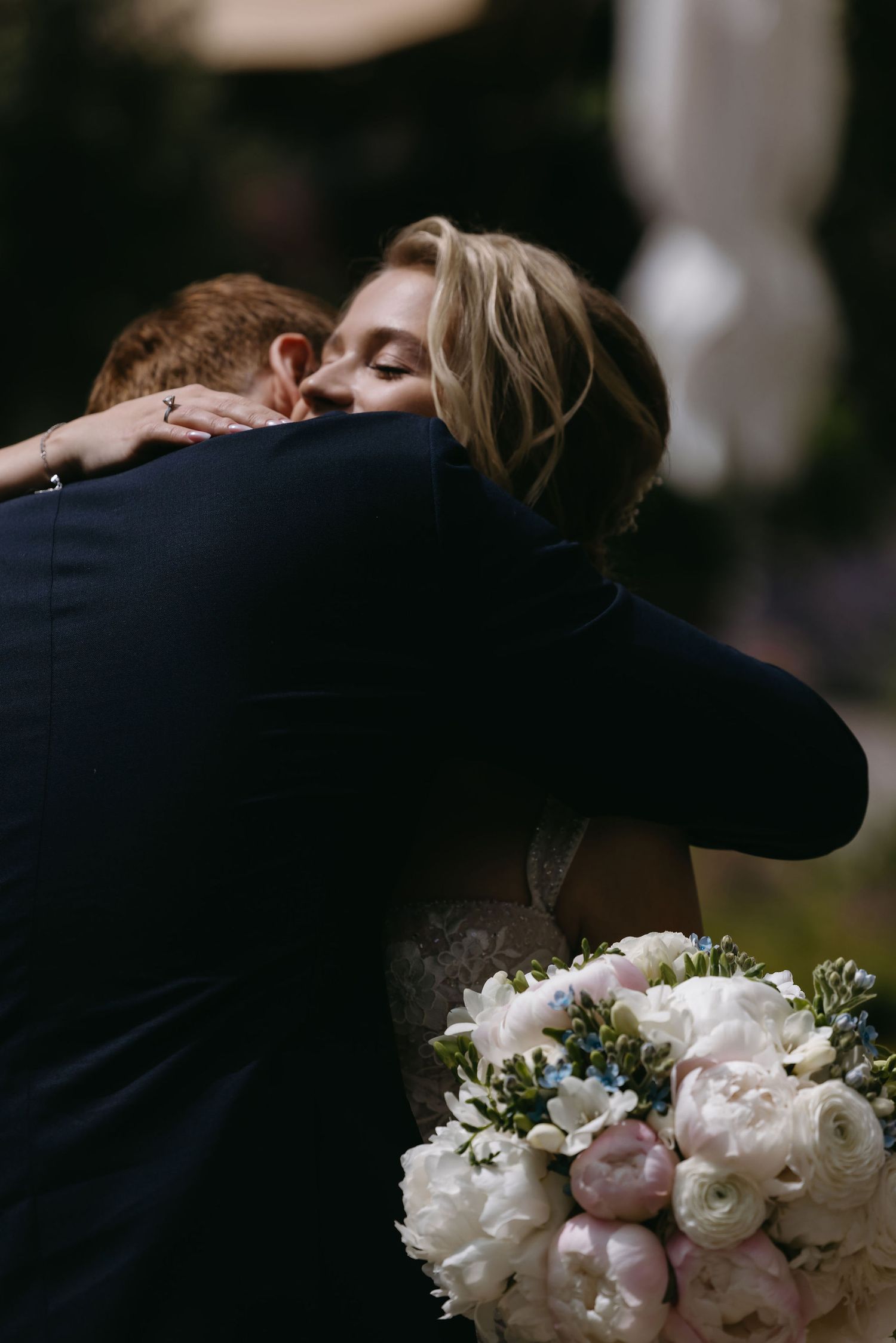 Two newlyweds embrace in a tender moment at their wedding with a white and pink rose bouquet visible in the foreground.