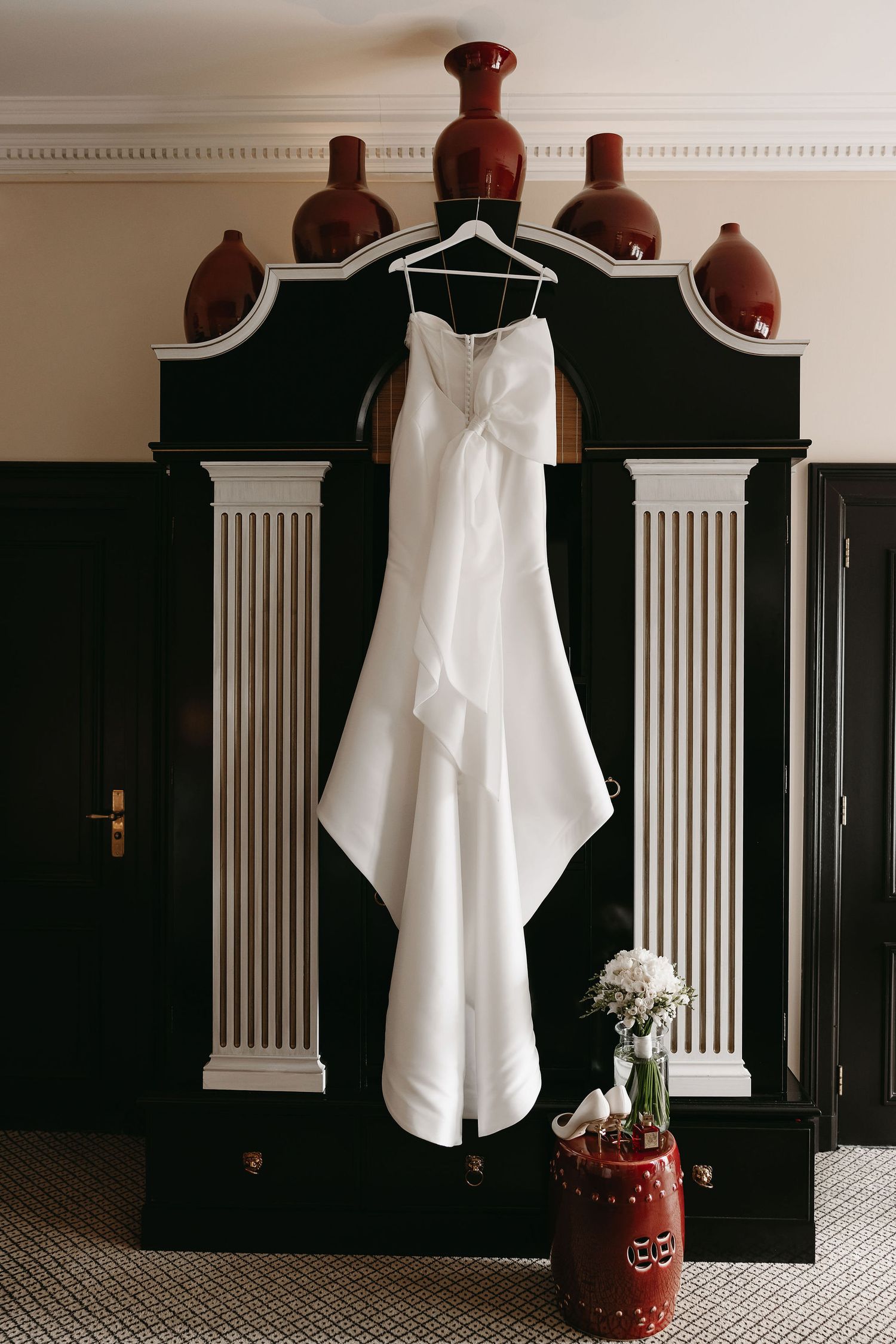 Elegant white wedding dress hanging on ornate dark wooden headboard with white trim and red vase with white flowers below.