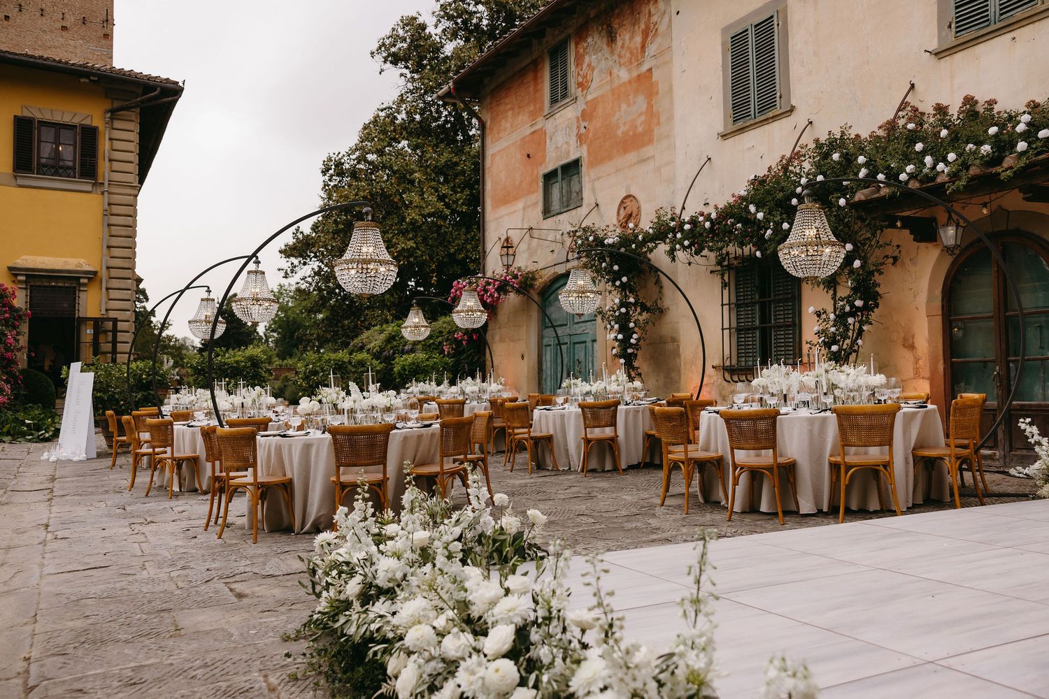 Elegant outdoor wedding reception setup with white floral arrangements at a rustic Italian villa courtyard.