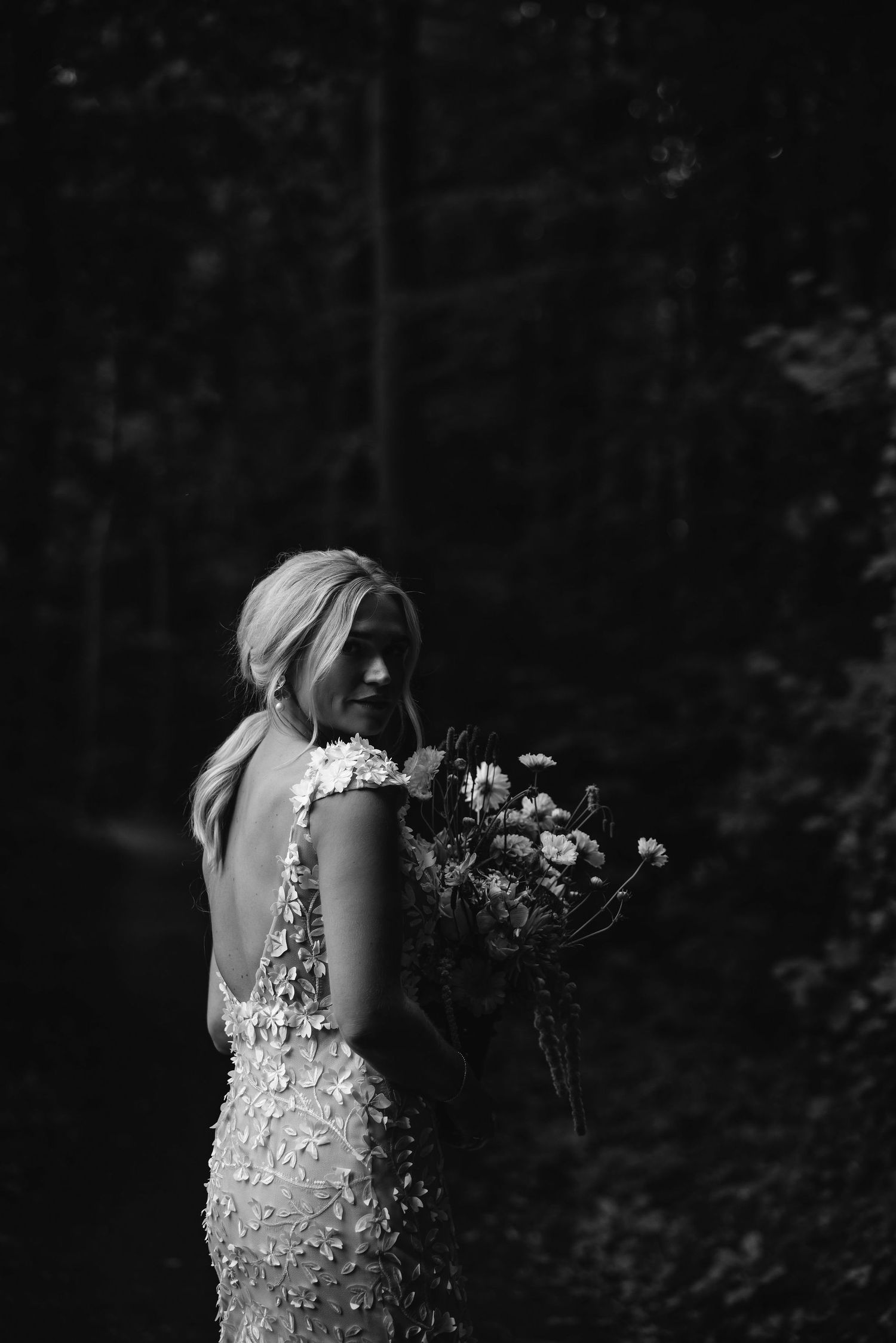 A bride in a detailed lace wedding dress holds a floral bouquet while looking over her shoulder in a dark outdoor setting.
