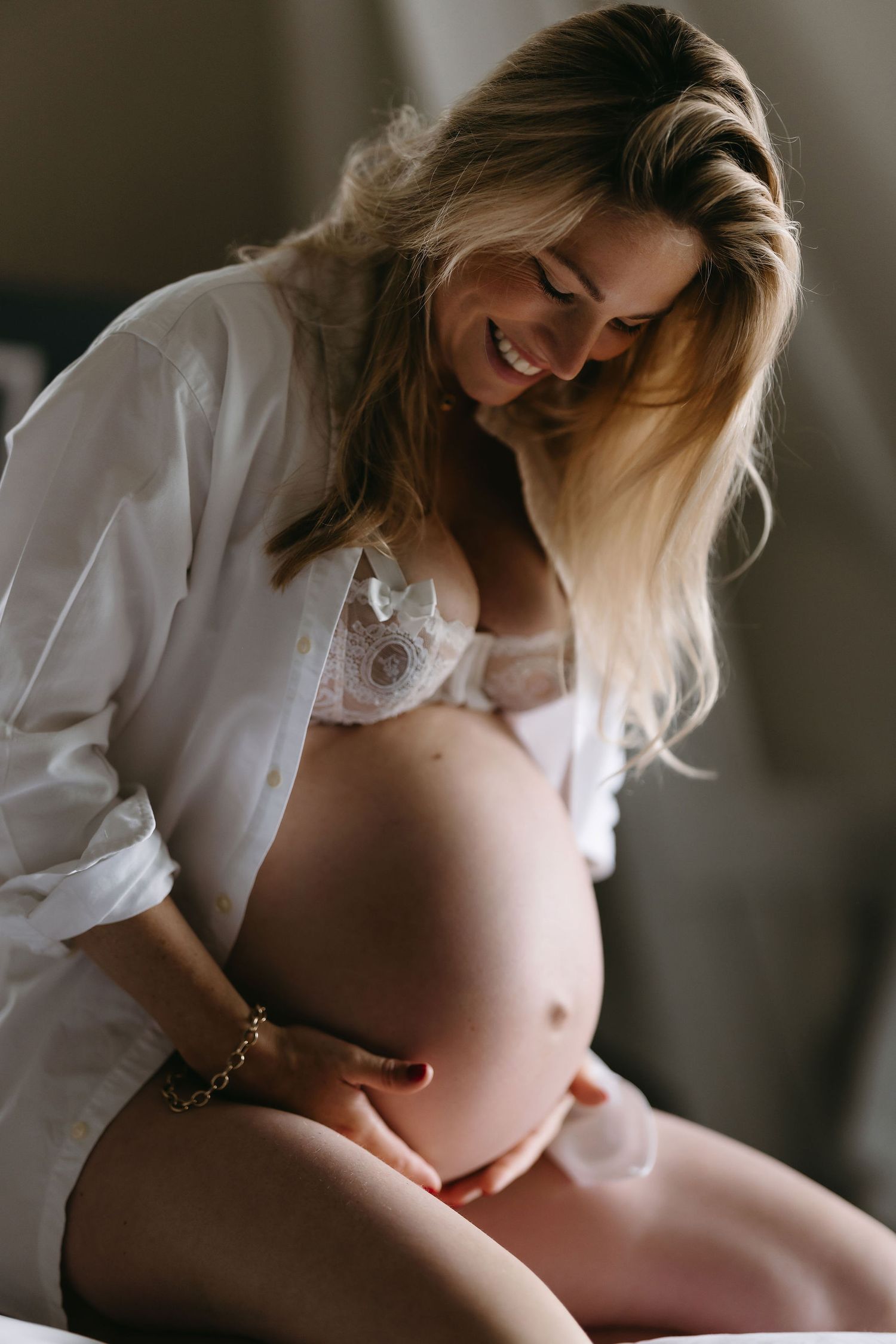 A tender maternity boudoir photo showing an expectant mother in white button-down shirt and lace bralette.