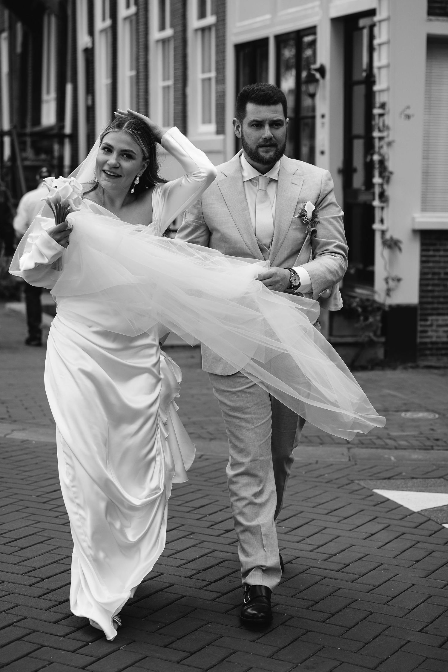 A couple dressed in white and grey run joyfully along a cobblestone street on their wedding day.