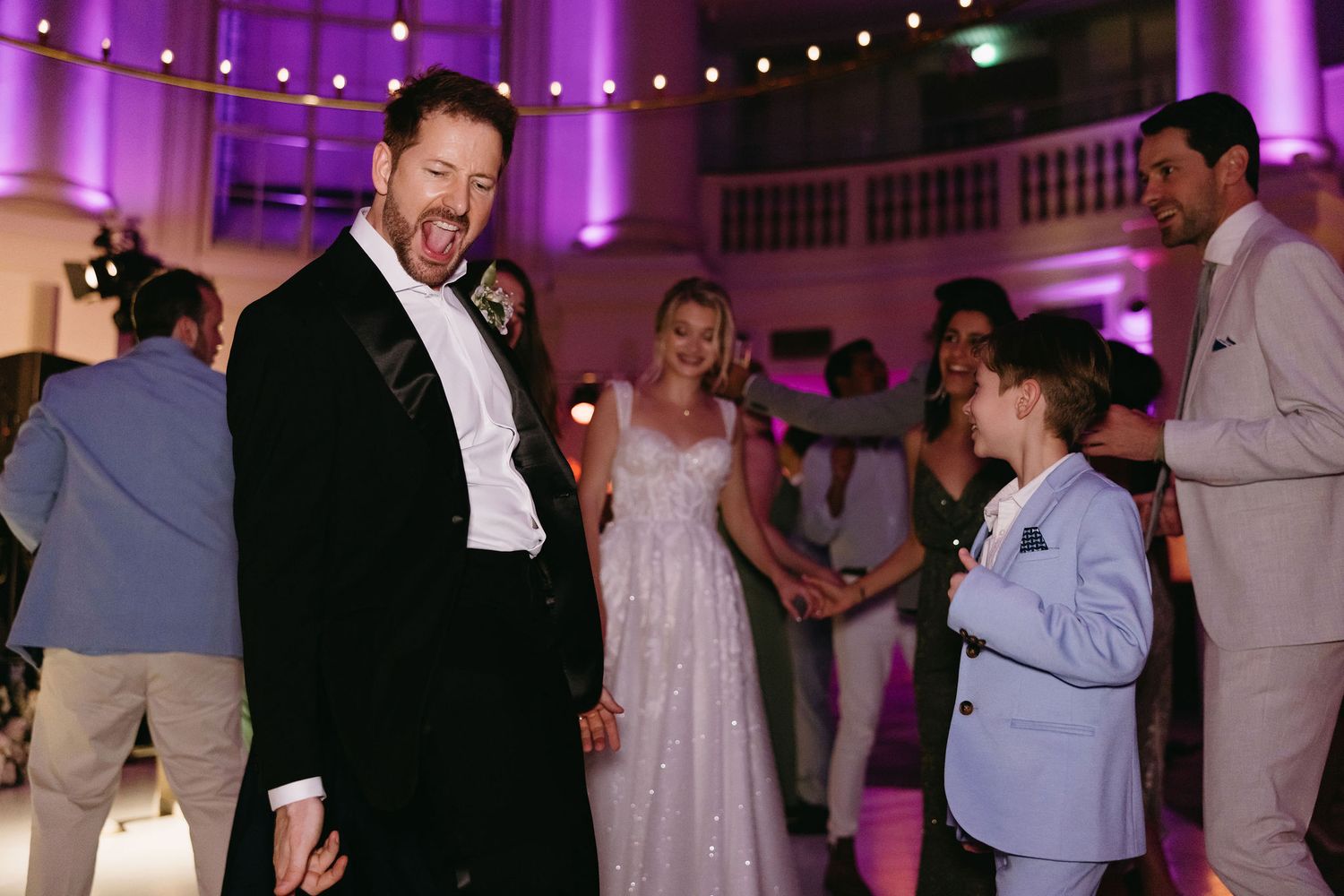Wedding guests dancing and celebrating in a purple-lit reception venue during an energetic party moment.