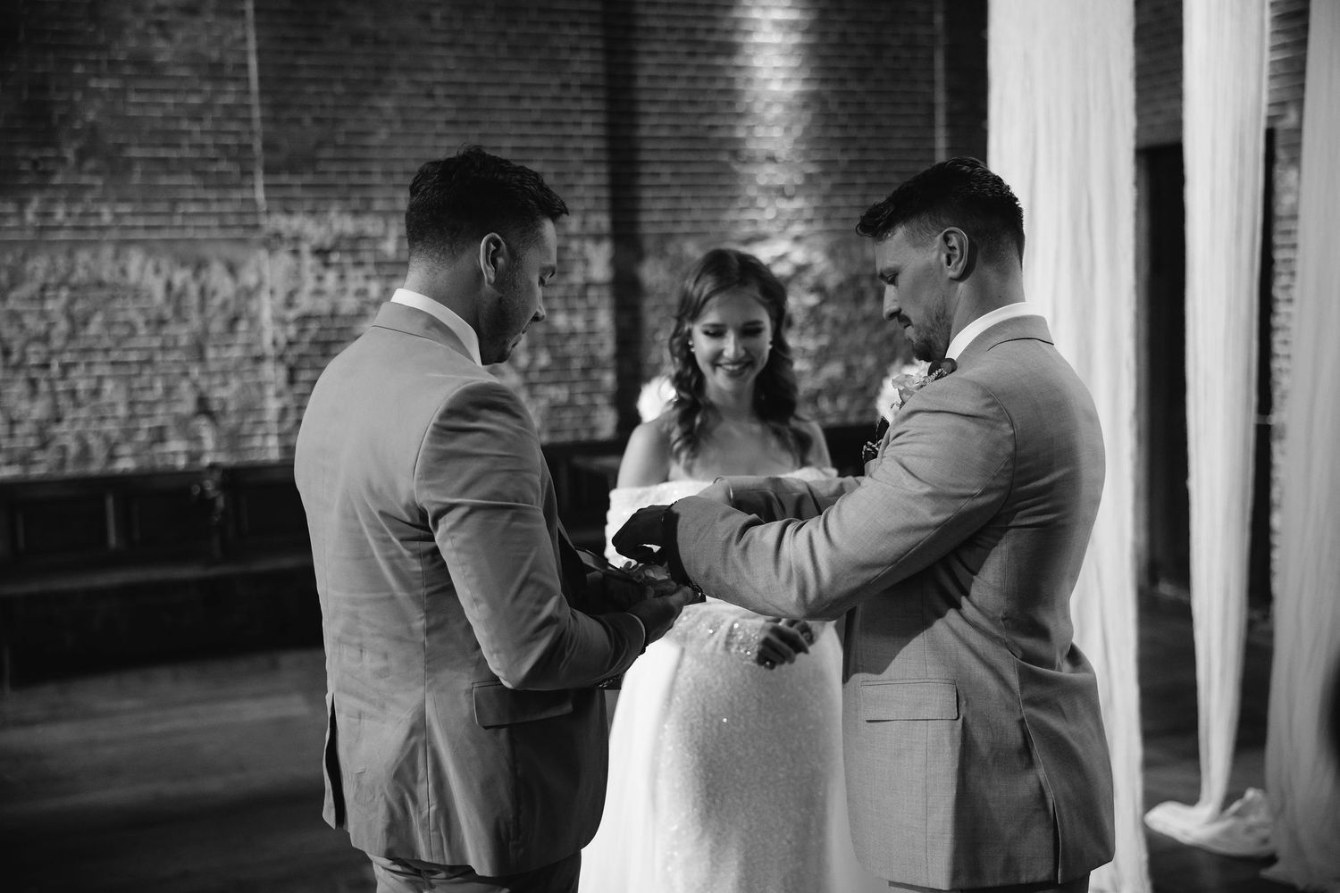 An intimate moment between a bride in white and two groomsmen in light colored suits at an indoor venue.