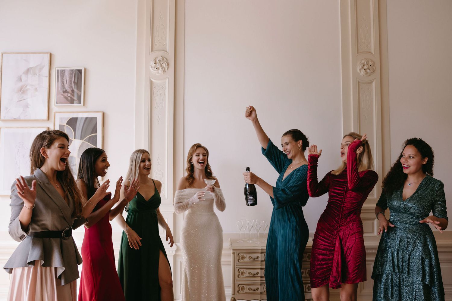Wedding party members in jewel-toned dresses posing against a white wall.