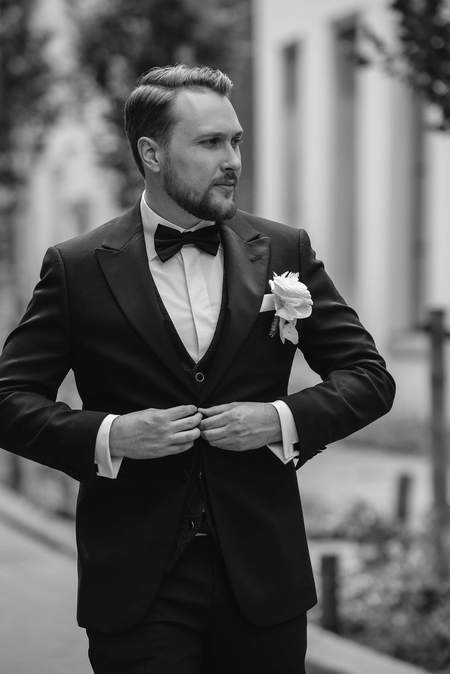 Classic black and white portrait of groom in tuxedo with boutonniere adjusting his jacket.