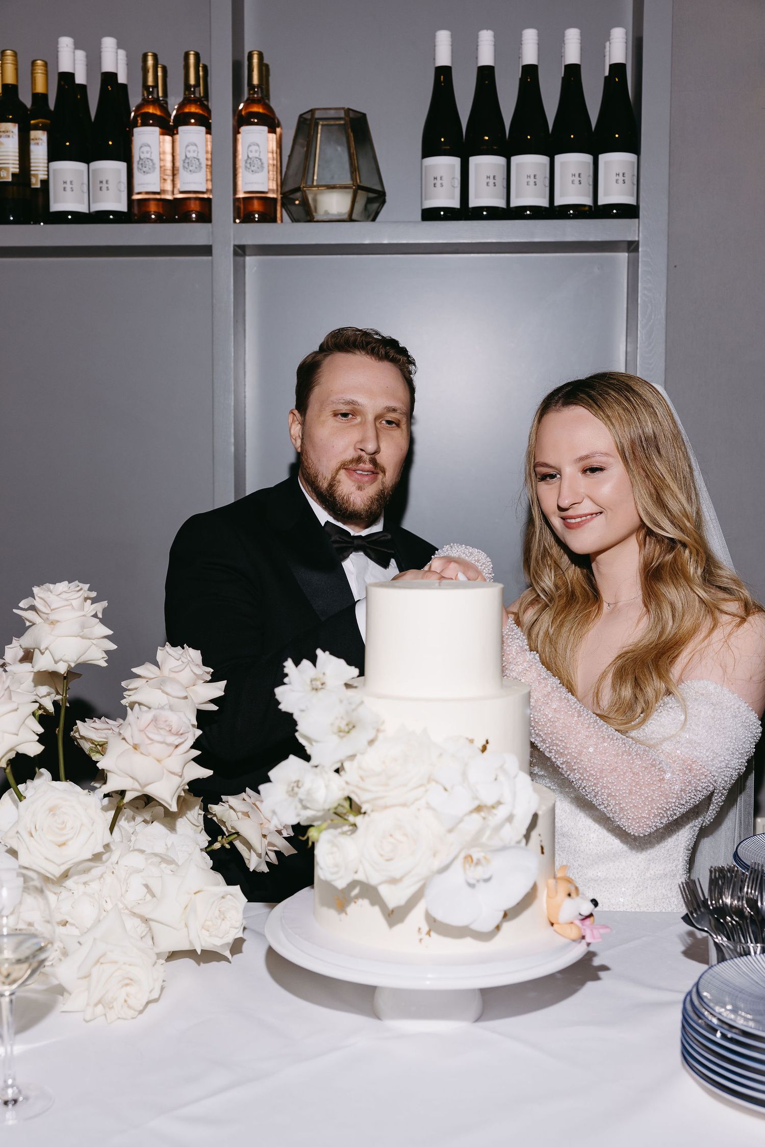 Newlyweds cutting their elegant white wedding cake surrounded by roses and wine bottles.