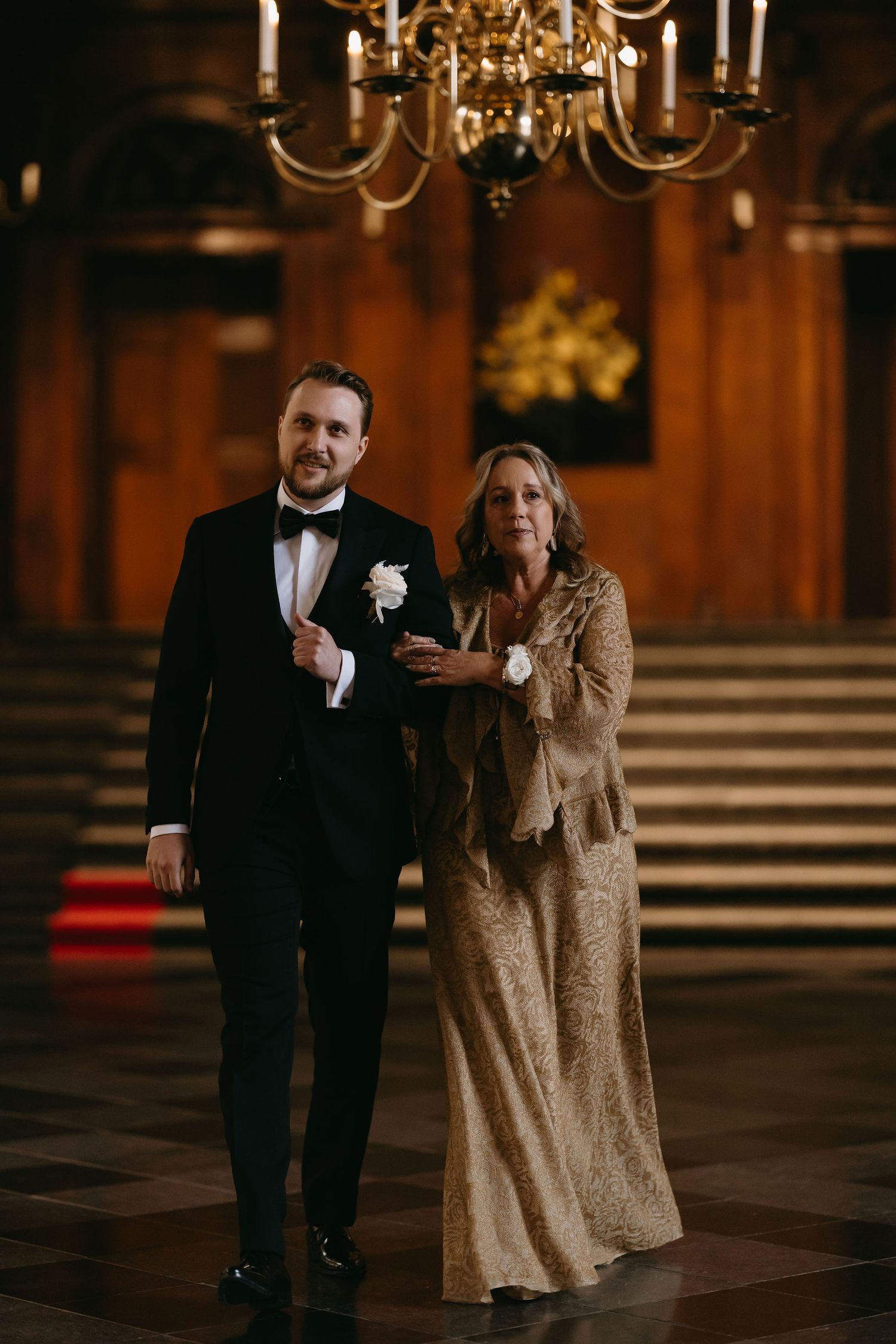 Two people in formal evening attire walk through an elegant wood-paneled room with a chandelier.