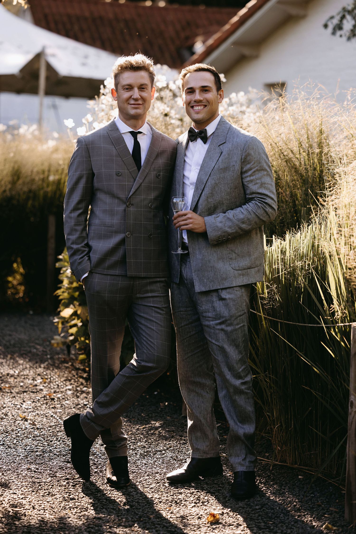 Two men in stylish gray suits stand together outdoors with tall grasses and sunlit backdrop.