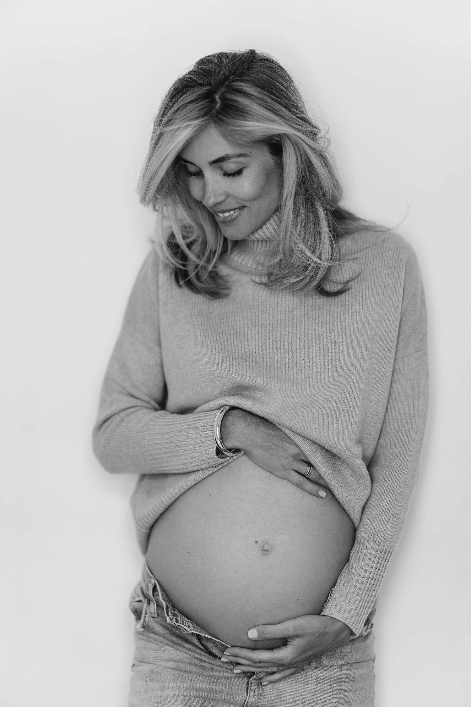 Maternity portraits in soft grey sweater showing baby bump against white background.