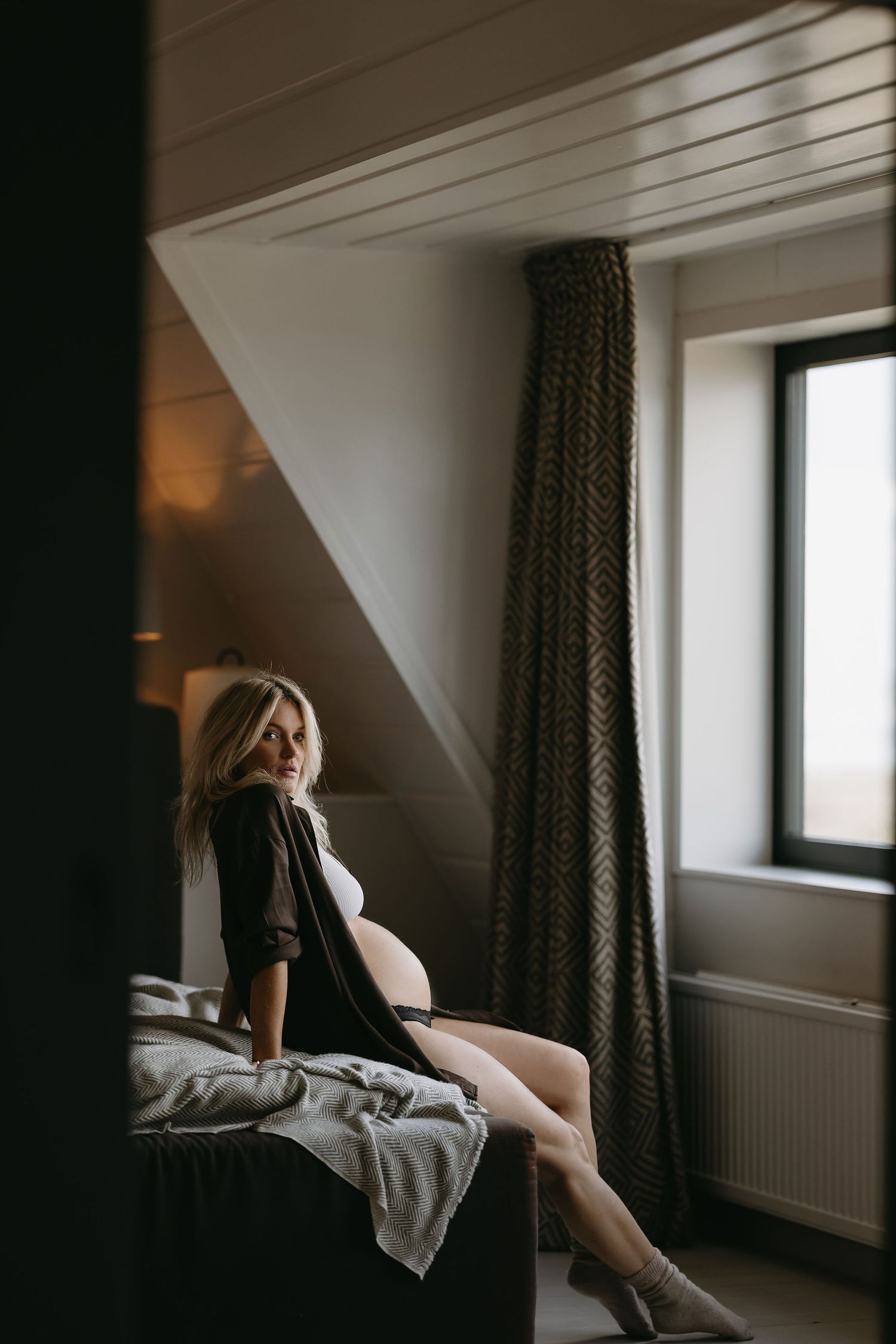 Moody boudoir photo of someone relaxing on a bed by a window in an attic bedroom.