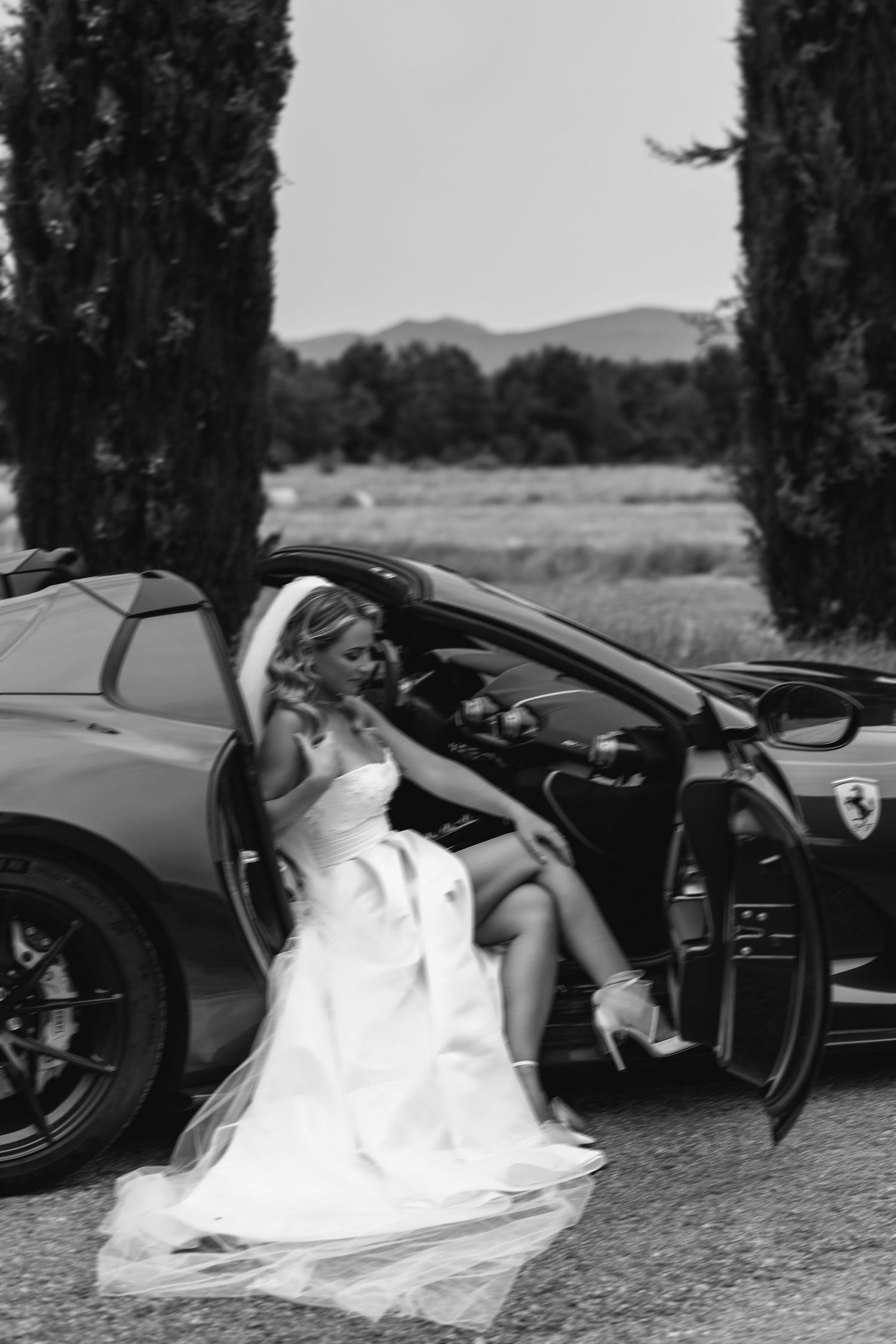 Bride in white gown poses elegantly in a Ferrari sports car with cypress trees in background.