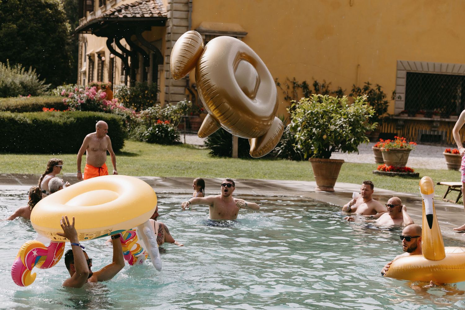 People playing in a pool with large inflatable donut and flamingo floats during a celebration.
