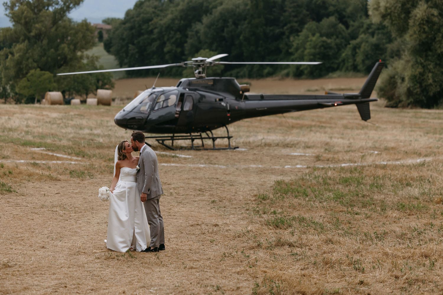 A wedding couple stands near a black helicopter in a grassy field.