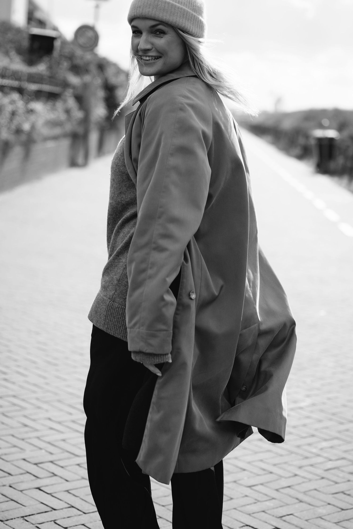 Black and white portrait of someone in a winter coat and beanie smiling on a cobblestone path.