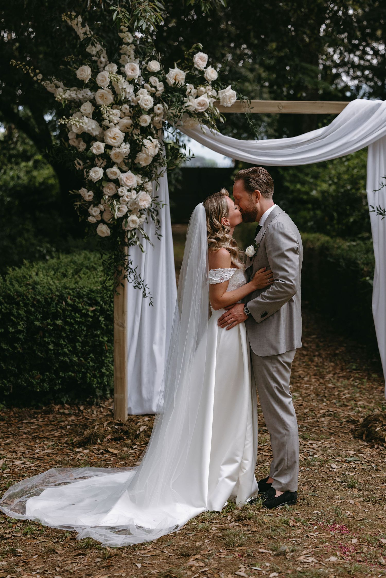 Romantic wedding ceremony under a white draped arch decorated with roses in a garden setting.