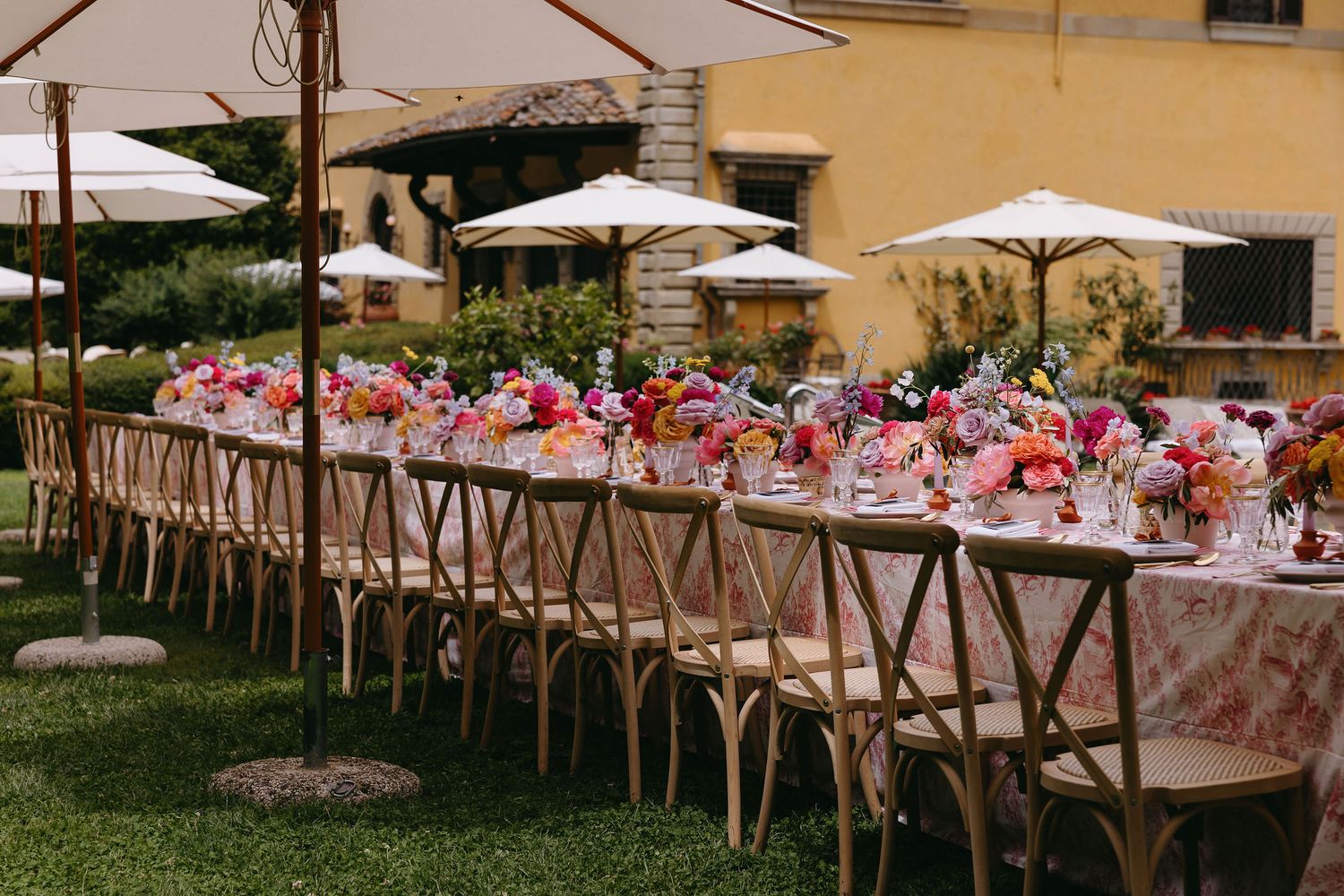 Long wedding reception table set with pink florals and cross-back chairs in an outdoor garden setting.