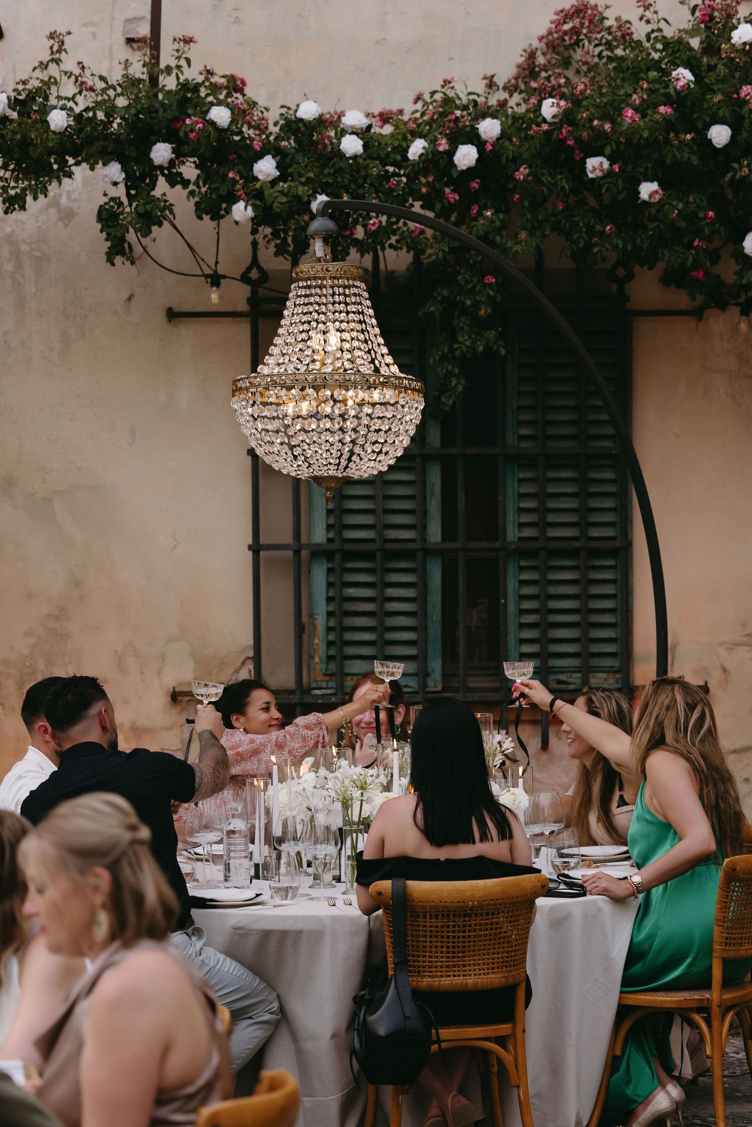 Crystal chandelier hangs over an outdoor dinner table with climbing roses.