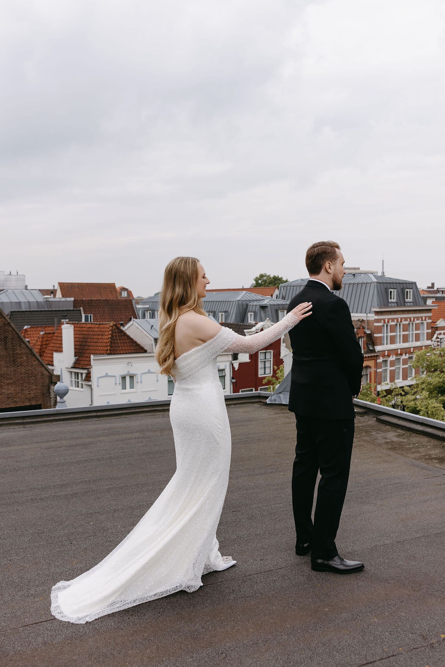 Couple in wedding attire pose playfully on urban rooftop with city buildings behind them.
