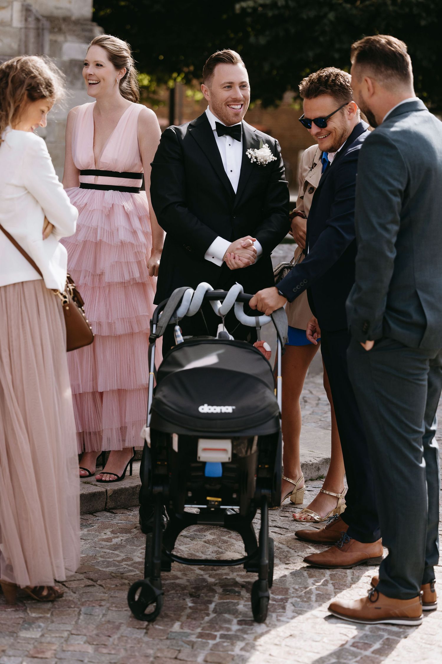 Wedding guests in formal attire gather around a baby stroller outdoors.