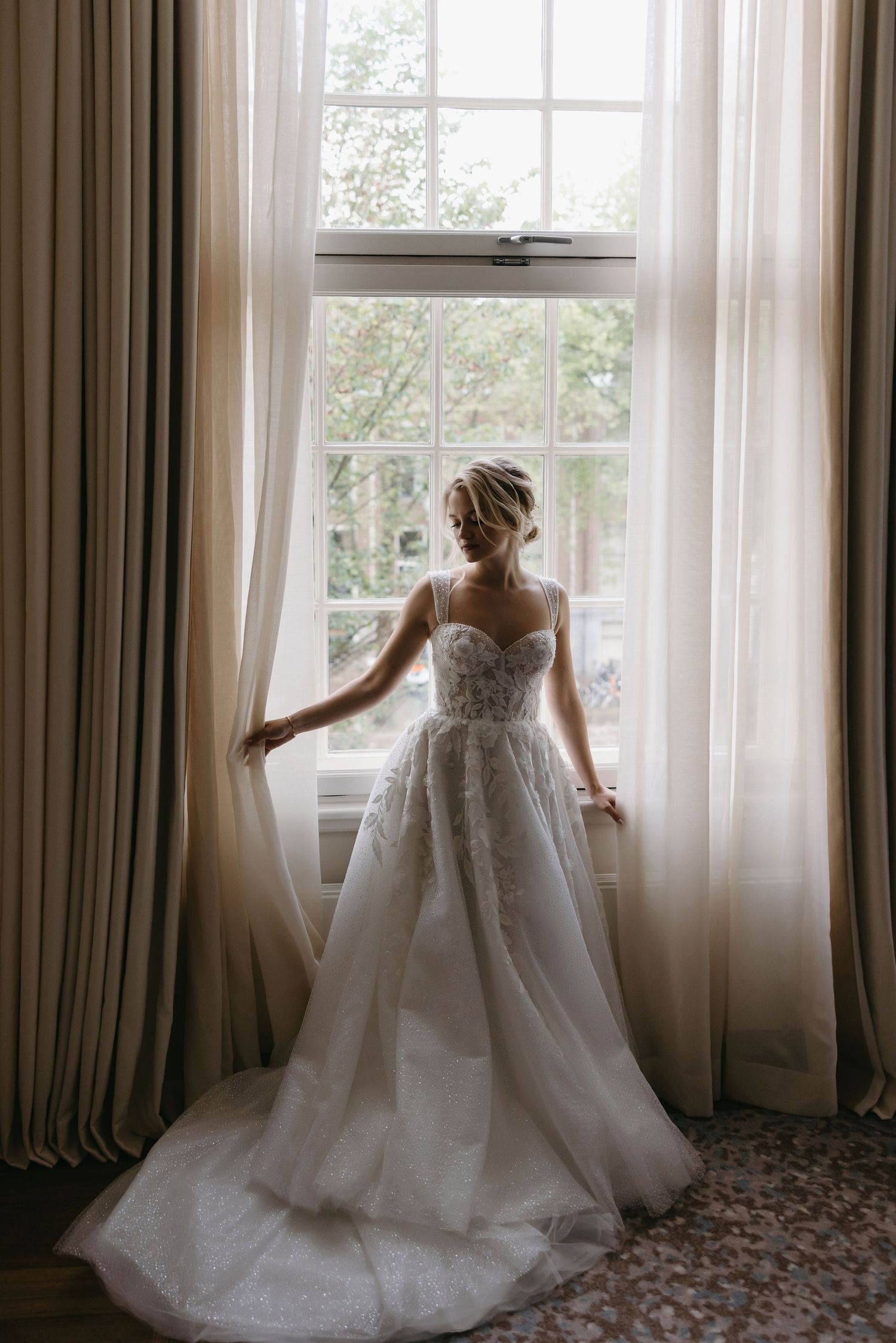 Sunlit bridal gown displayed against tall windows in an elegant room with flowing curtains.
