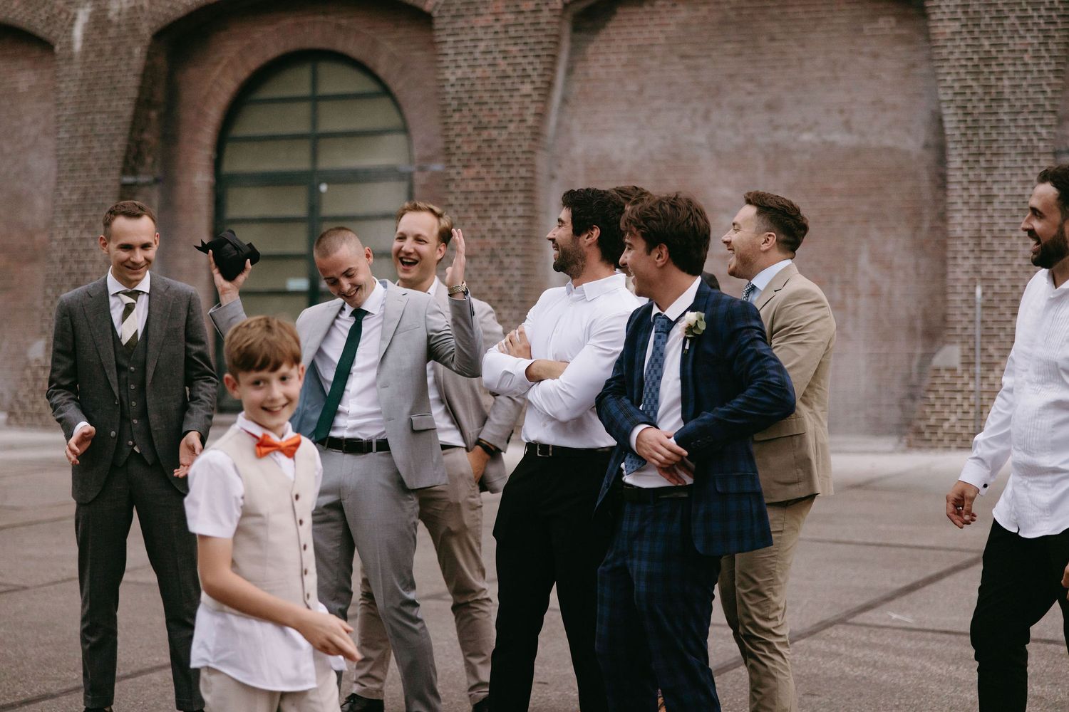Wedding party members gather outside a brick building for celebratory group photos.
