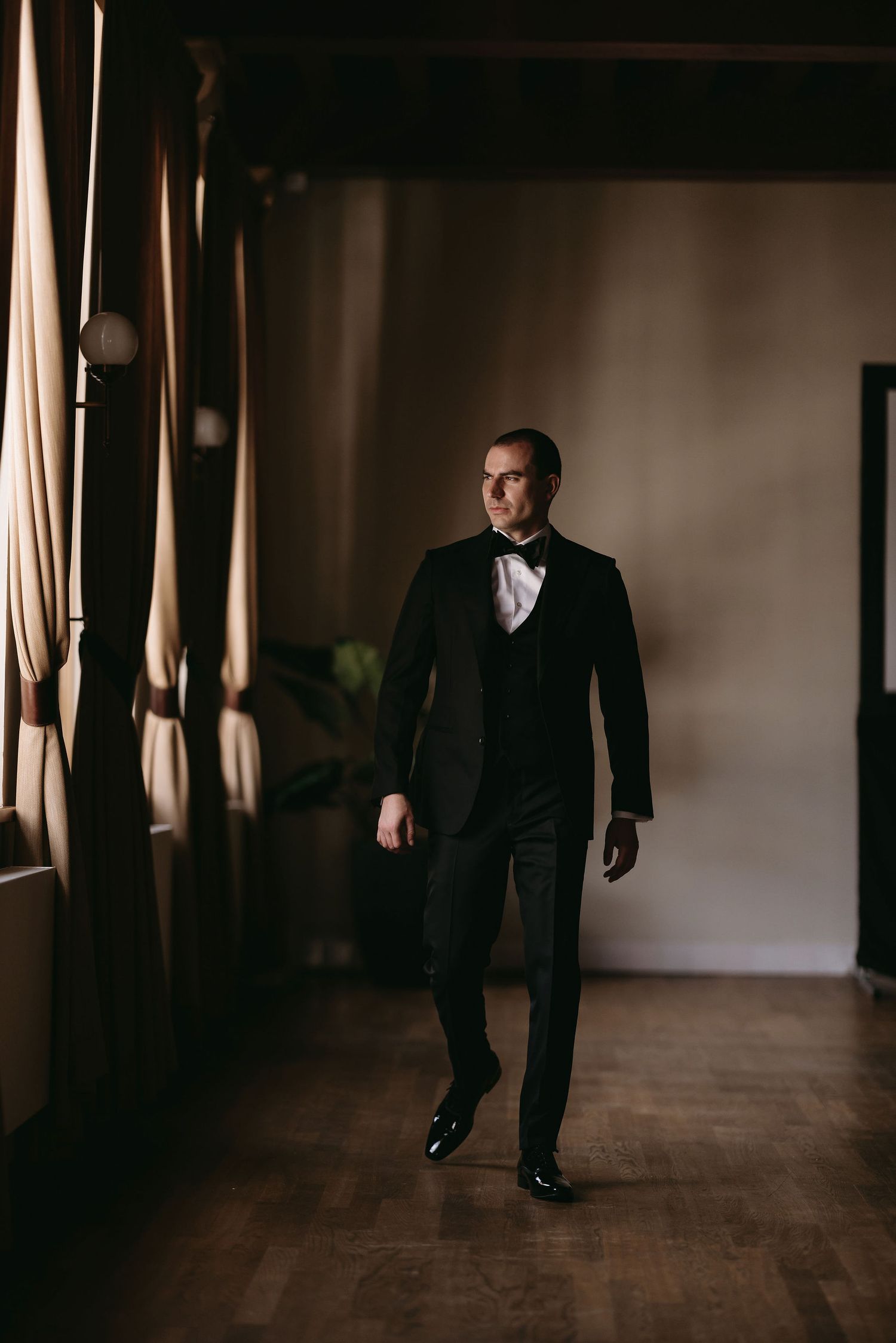 A person in an elegant black tuxedo and bow tie walks gracefully across a hardwood floor in a moody interior setting.