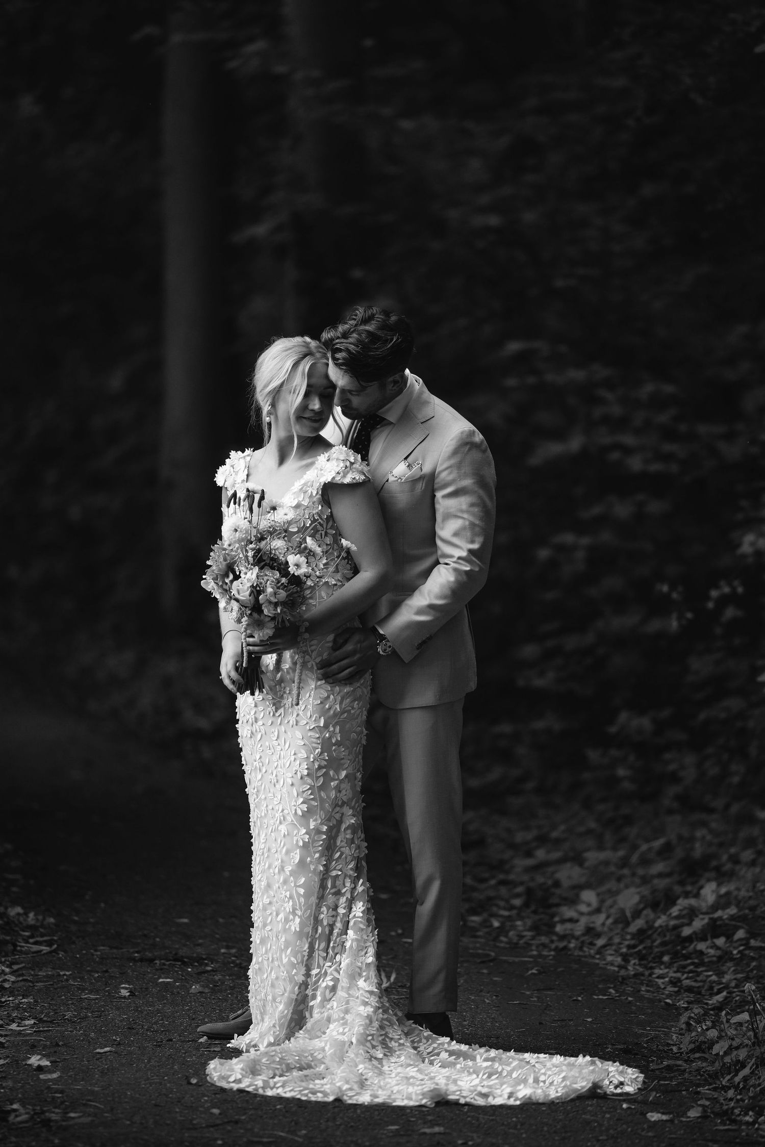 Romantic black and white portrait of a couple in formal attire against a forest backdrop.