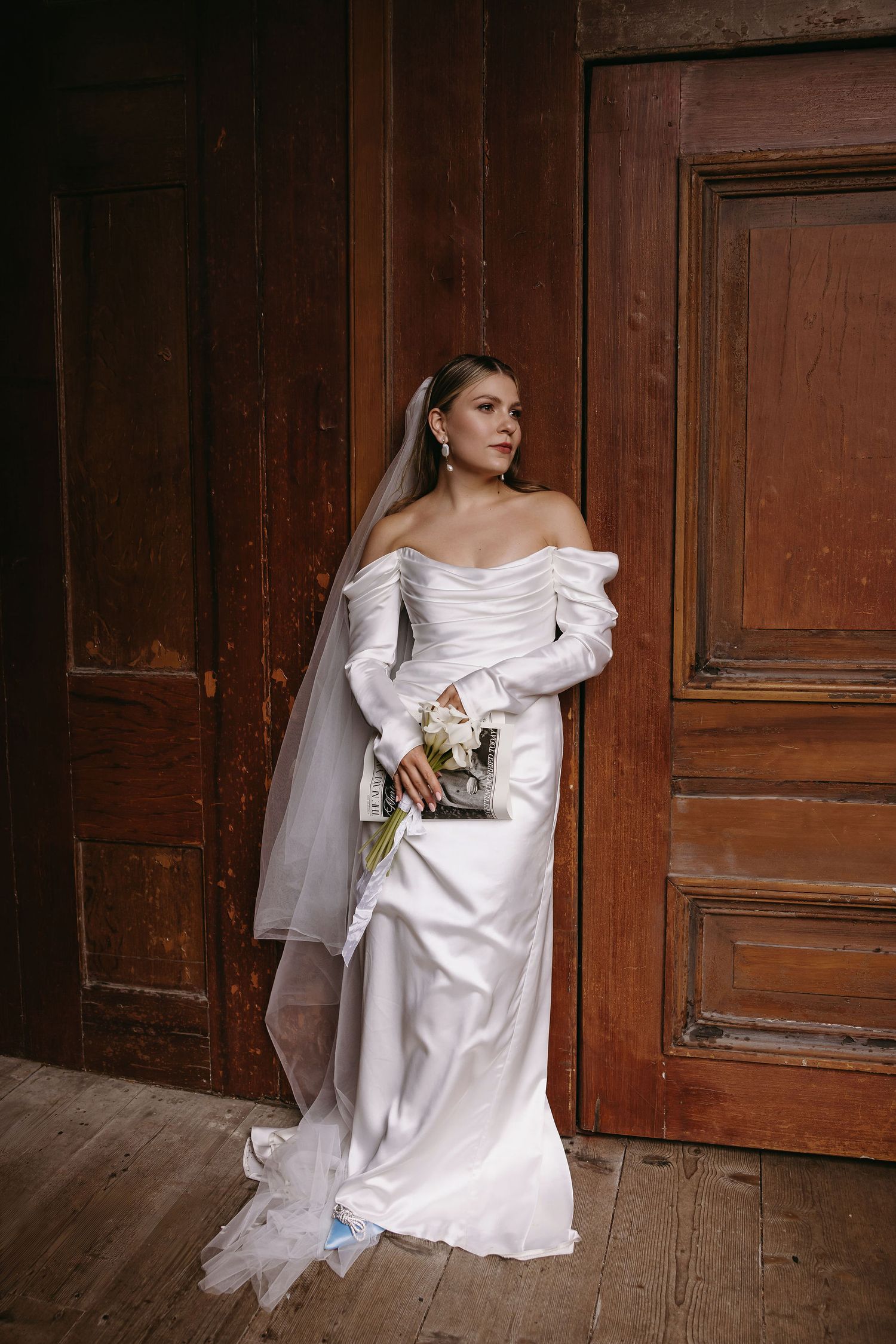 Bride in elegant white dress sitting against vintage wooden doors.