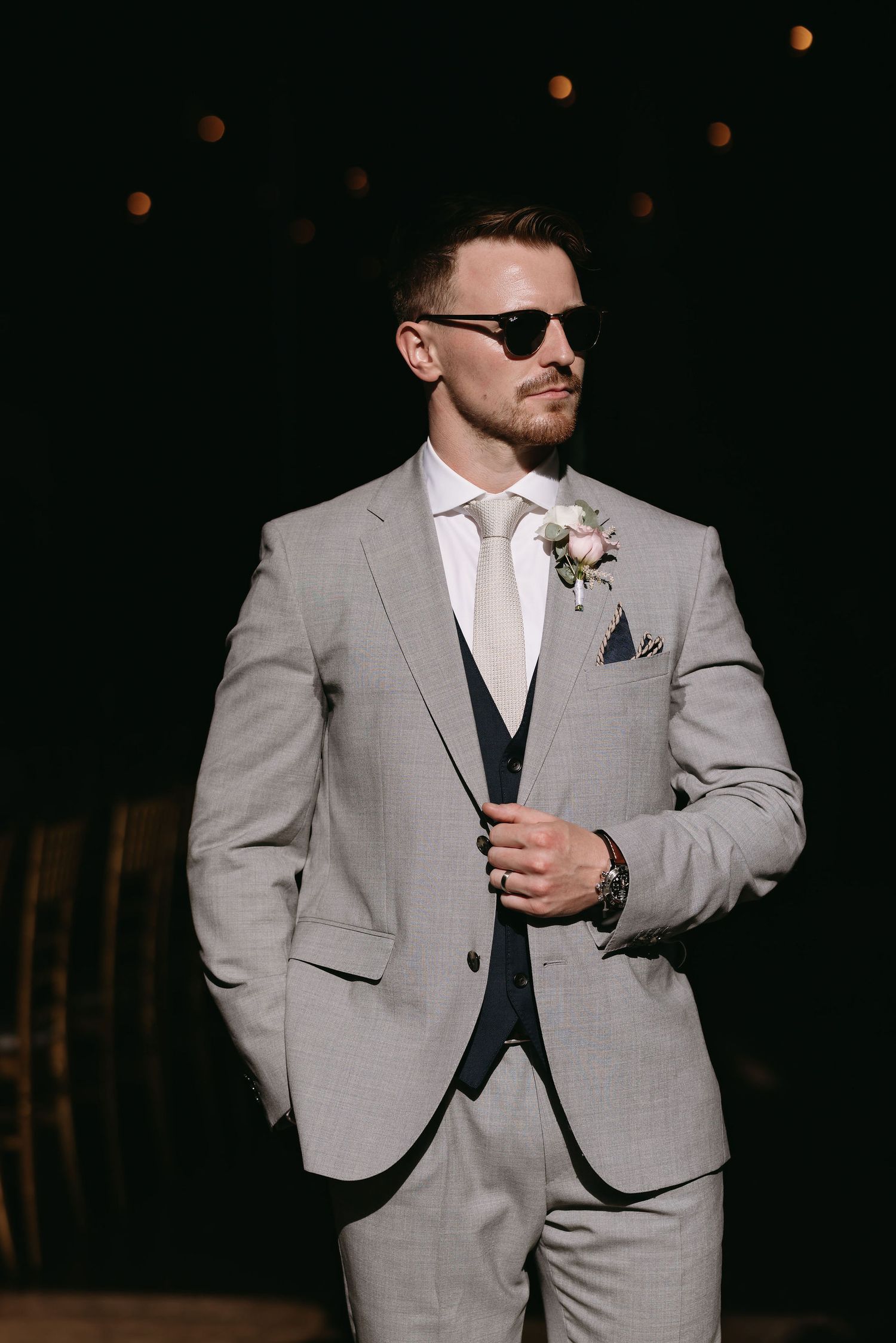 A groom in a light grey suit and white tie stands confidently in dramatic lighting.
