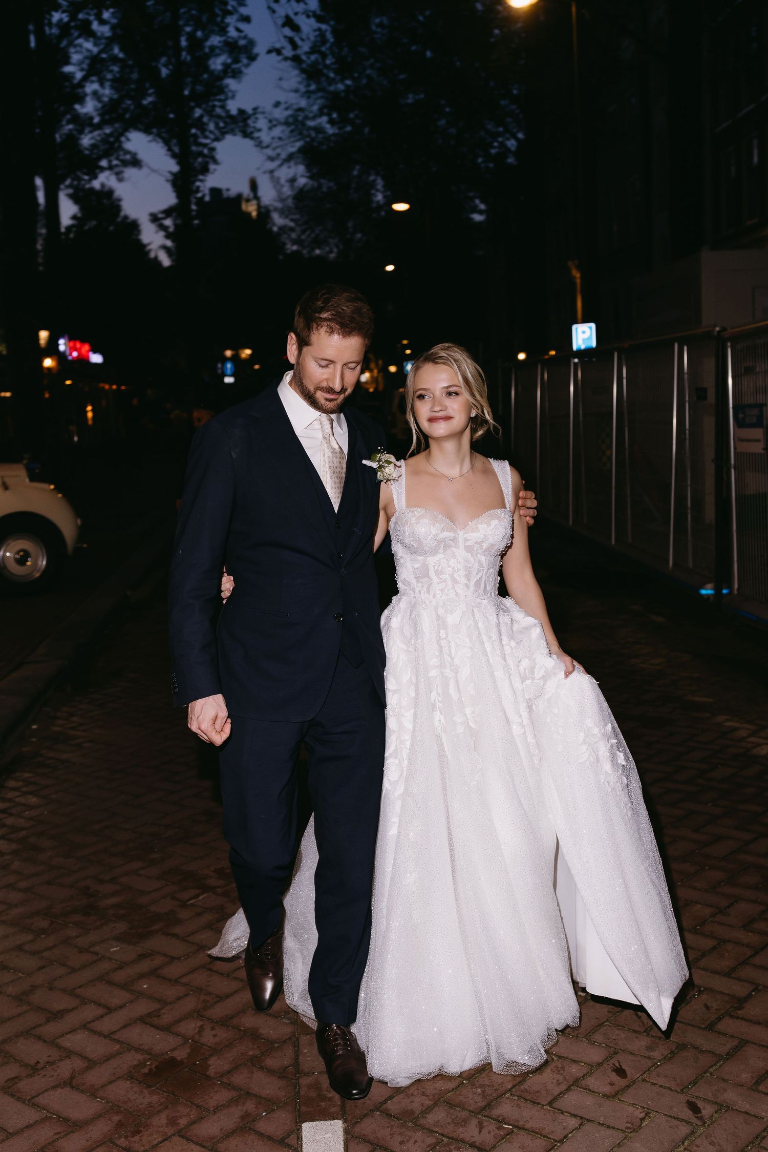 Couple walking together at night on brick pathway in formal attire.