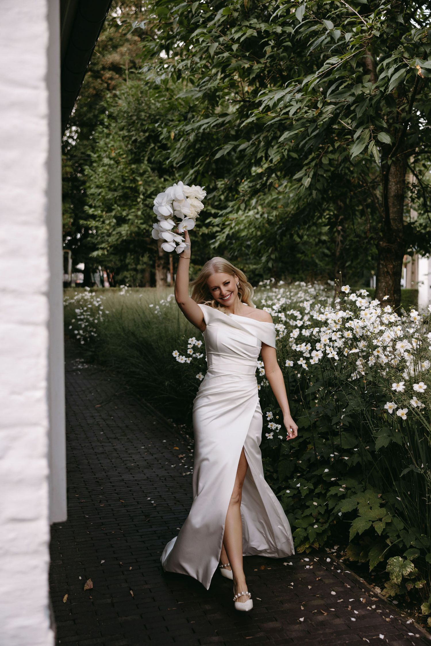 A bride in sleek white dress holds bouquet aloft while walking through garden pathway.