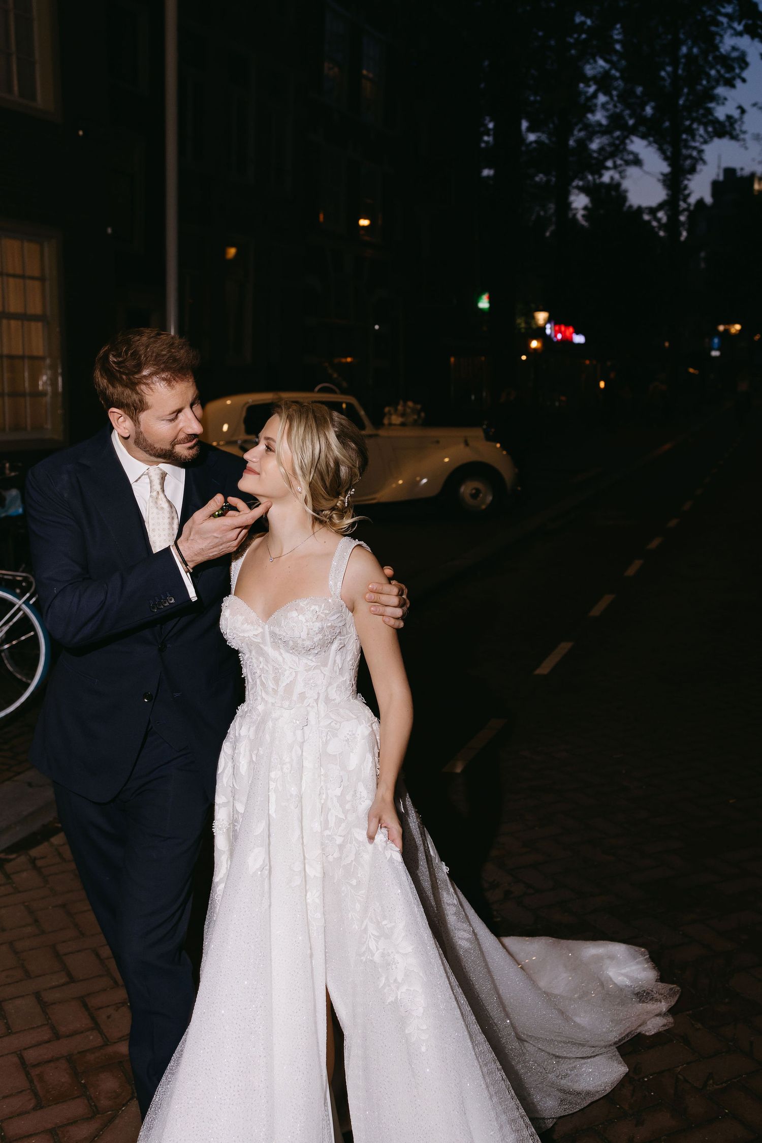 A couple shares a romantic nighttime moment on a cobblestone street in formal wedding attire.