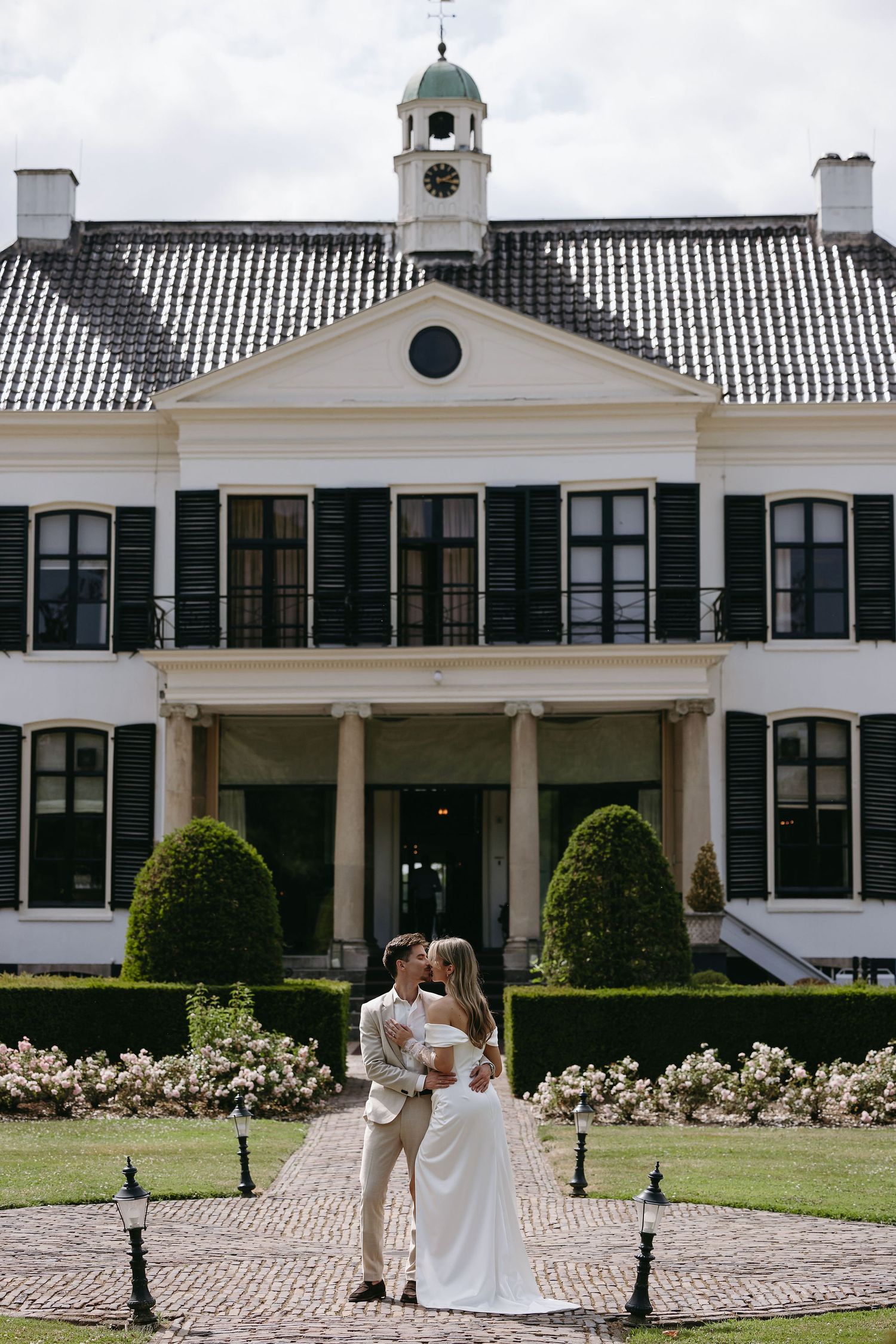 A wedding scene in front of a grand white colonial mansion with columns and landscaped gardens.