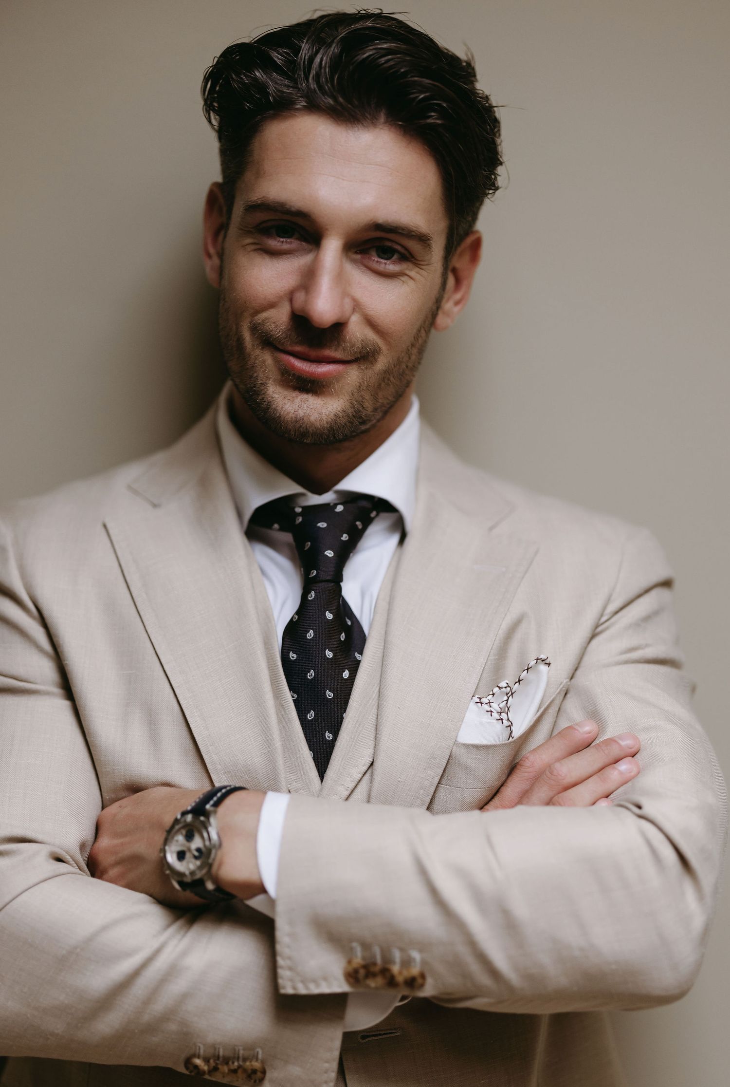 Stylishly dressed man in beige suit and polka dot tie poses confidently with crossed arms and a wristwatch.